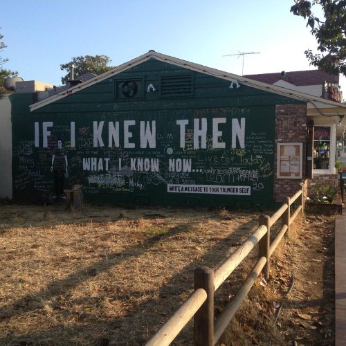 A green wall with chalk messages, including "If I knew then what I know now," near a small building and a field.