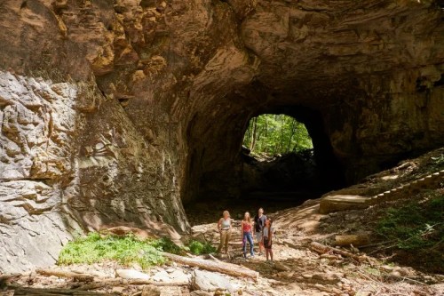 Three people explore a large cave with rocky walls and greenery in the background. Sunlight filters through the entrance.