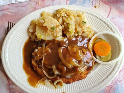 A plate of food featuring a breaded cutlet topped with gravy, onions, and sides of mashed potatoes and vegetables.