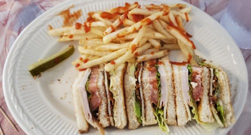 A plate with a club sandwich, layered with lettuce, tomato, and turkey, accompanied by fries and a pickle.