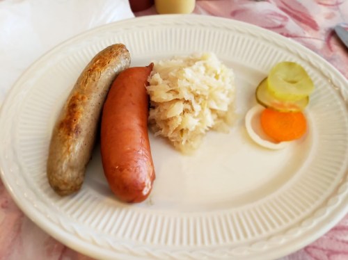 A plate with two sausages, sauerkraut, and pickled vegetables including carrots and cucumbers.