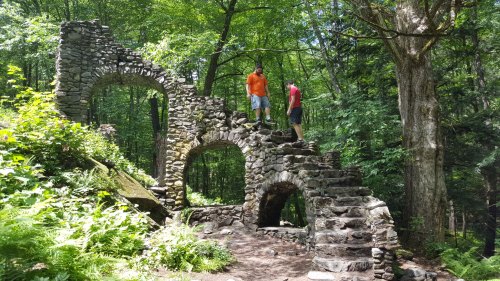 Two people stand on a stone structure with arches, surrounded by lush greenery in a forested area.