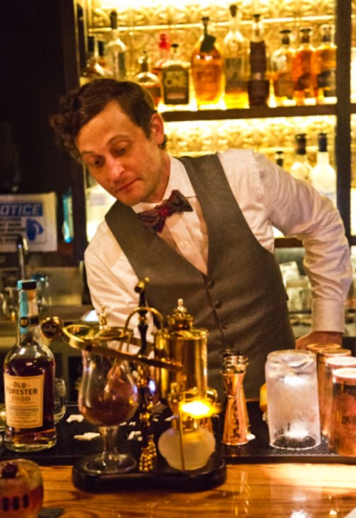 A bartender in a vest and bow tie prepares a drink at a well-stocked bar with various bottles in the background.