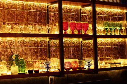 A display of colorful glassware on illuminated shelves against a decorative wall.
