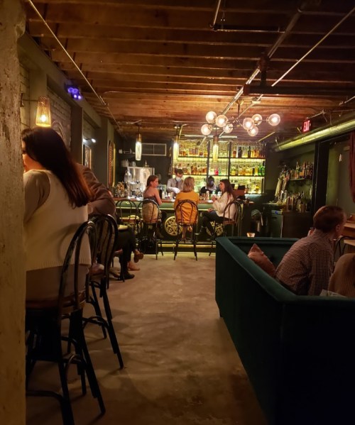 A cozy bar scene with patrons seated at the counter and tables, warm lighting, and a stylish decor.