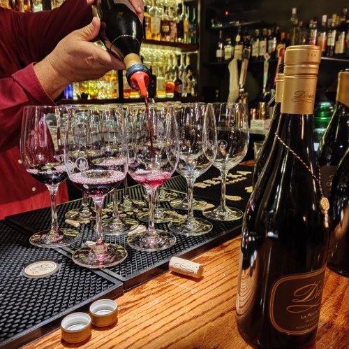 A bartender pours red wine into several glasses at a bar, with a bottle of wine and corks on the counter.