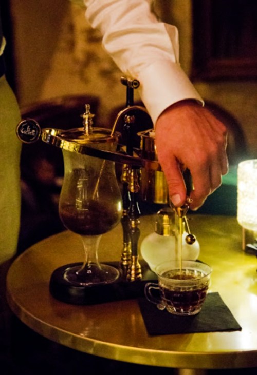 A hand pours coffee from a stylish brass coffee maker into a small glass cup on a table.