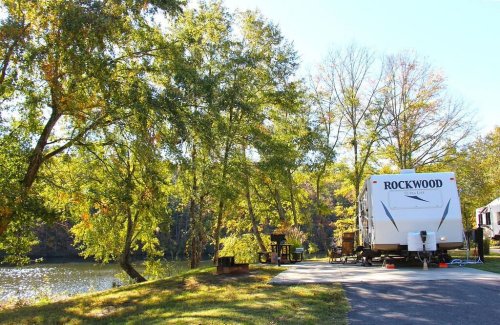 A camper parked by a serene lake, surrounded by trees and autumn foliage.