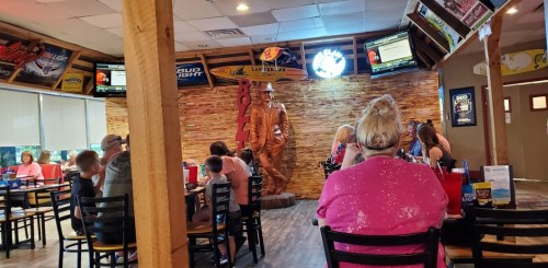 A restaurant interior with patrons seated, featuring a wooden wall and a statue of a man in a cowboy hat.