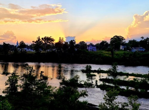 Sunset over a river, with colorful clouds and reflections on the water, surrounded by trees and distant houses.