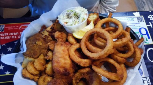 A platter of fried seafood, including fish and shrimp, served with onion rings, coleslaw, and a lemon wedge.