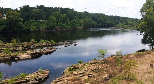 A serene river scene with rocky banks, lush greenery, and calm waters under a partly cloudy sky.
