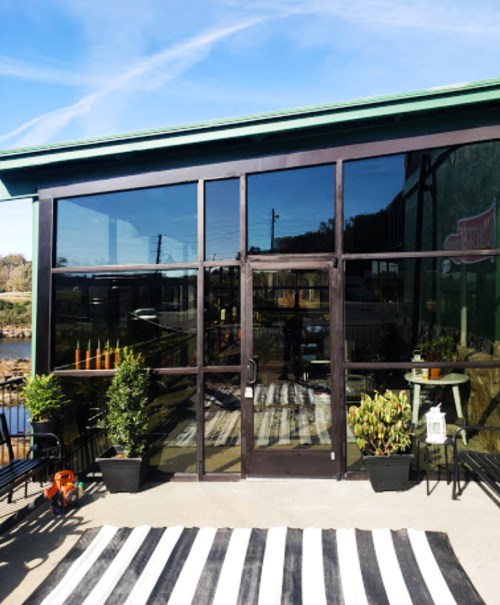 A modern glass-fronted building with a green roof, featuring a striped outdoor rug and potted plants.