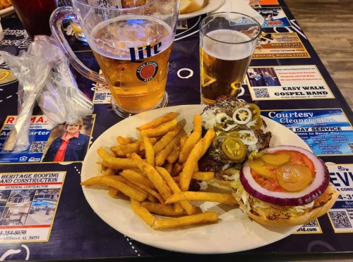 A plate with a burger topped with jalapeños and onion, served with fries and two glasses of beer on a table.