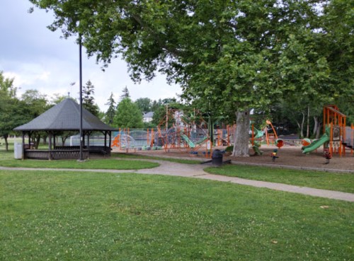A park scene featuring a gazebo, playground equipment, and green grass under a cloudy sky.