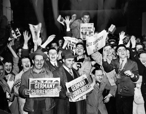 Crowd celebrating Germany's surrender, holding newspapers with headlines about the surrender, joyful expressions all around.