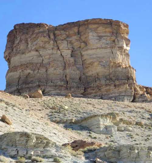 A tall, rocky cliff rises against a clear blue sky, surrounded by a dry, sandy landscape.