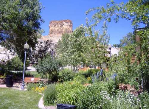 A lush garden with trees and flowers, set against a rocky cliff under a clear blue sky. Benches are visible in the background.