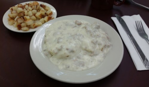 A plate of biscuits and gravy with a side of golden-brown diced potatoes on a table. Fork and knife beside the plate.