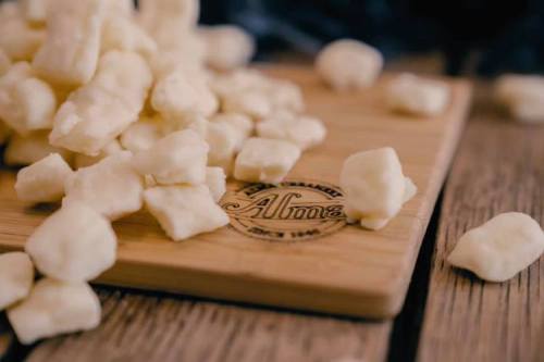 A wooden board with the engraved name "Alma" and scattered pieces of fresh cheese curds.