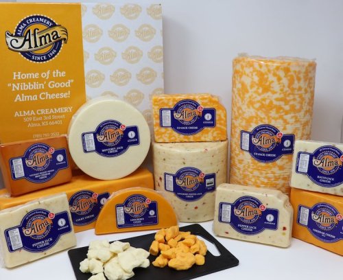 A variety of cheeses from Alma Creamery, including blocks and slices, displayed with a black platter of cheese snacks.