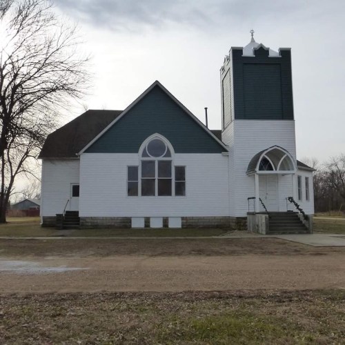 A white church with green accents, featuring a tall steeple and large windows, set against a cloudy sky.