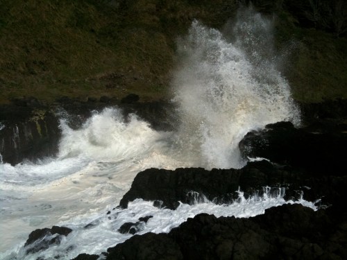 Waves crashing against rocky shore, creating a spray of water against a backdrop of green hills.