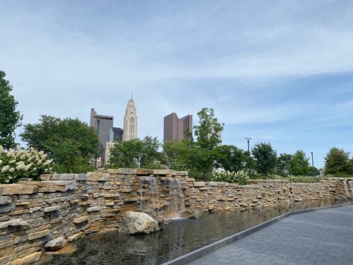 A serene urban scene featuring a stone waterfall and pond, with city skyscrapers in the background under a blue sky.