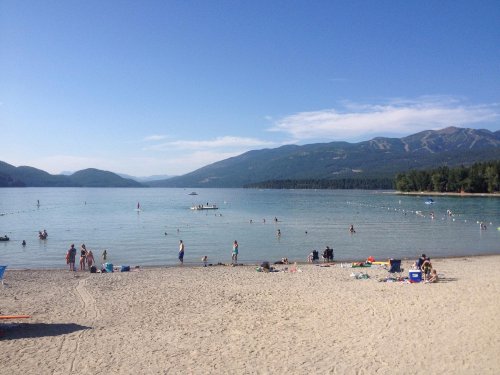 A sandy beach by a lake with people swimming and relaxing, surrounded by mountains and clear blue skies.