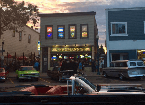 A vintage car parked in front of Neumann's bar, with colorful neon signs and a sunset in the background.