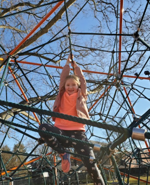A smiling child hangs from a climbing structure at a playground on a sunny day.