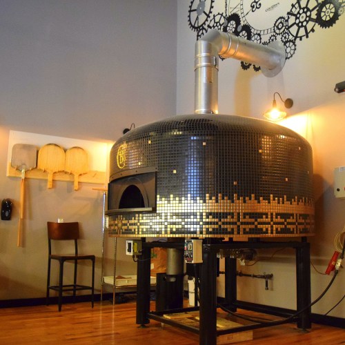 A large, round pizza oven with a mosaic design, situated in a cozy restaurant setting with wooden tools on the wall.