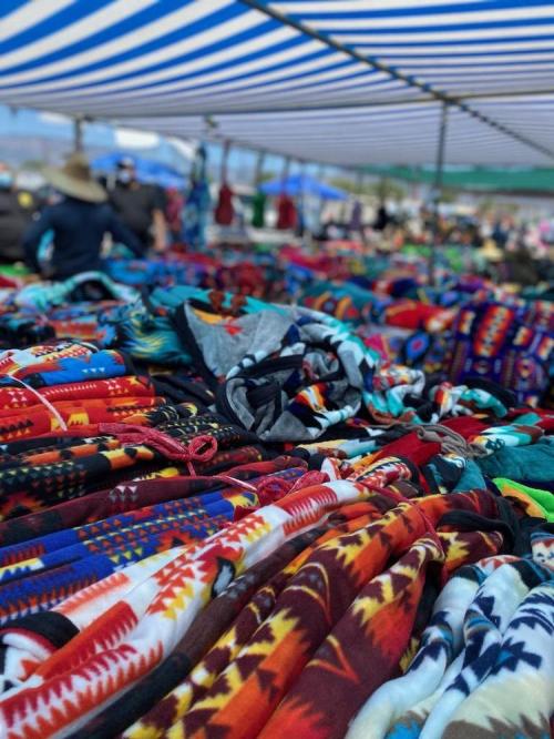 A vibrant market scene featuring a variety of colorful patterned blankets and textiles displayed under a striped canopy.