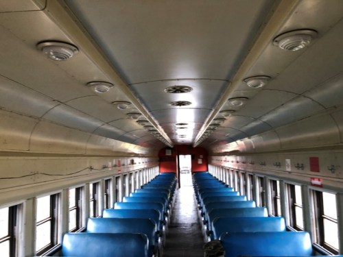 Interior of an empty train car with blue seats and overhead lights, leading to a red wall at the far end.