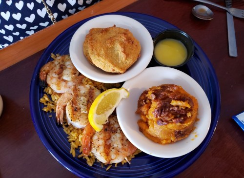 A plate featuring grilled shrimp with rice, cornbread, and a side of sweet potato with toppings.