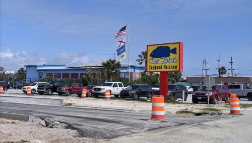 A seafood restaurant with a colorful sign, surrounded by parked cars and construction barriers under a clear sky.