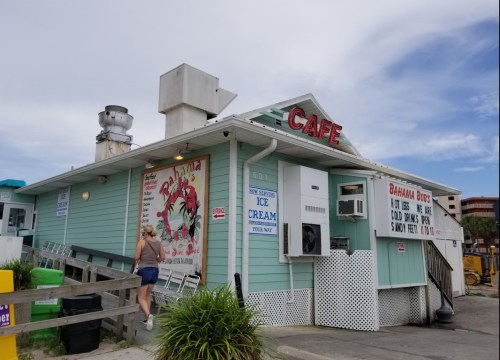 A turquoise café with a sign reading "Cafe" and a colorful mural, surrounded by greenery and outdoor seating.