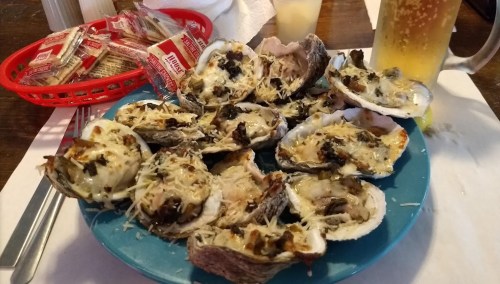 A blue plate filled with baked oysters topped with cheese, alongside a basket of crackers and a drink.