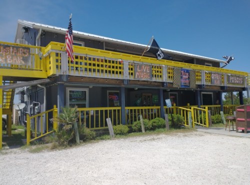 A colorful bar with yellow railings, flags, and signs for live music, set against a clear blue sky.