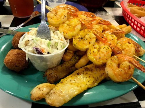 A colorful plate featuring grilled shrimp skewers, fish fillets, coleslaw, and hushpuppies on a teal platter.