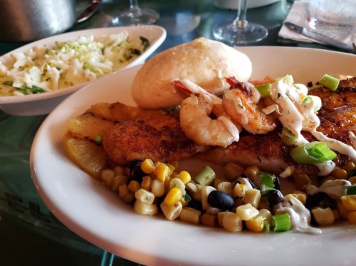 A plate of grilled fish topped with shrimp and green onions, served with corn and black bean salad and a roll.
