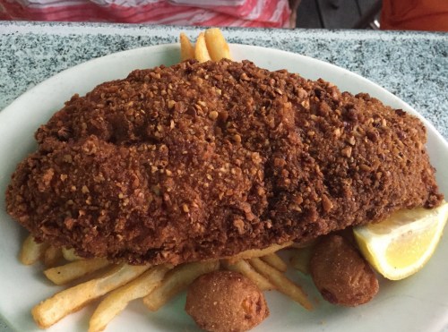 A large, golden-brown fried fish fillet on a plate with French fries and two hushpuppies, garnished with a lemon wedge.