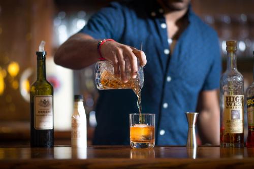 A bartender pours whiskey from a crystal decanter into a glass with ice, surrounded by various liquor bottles.