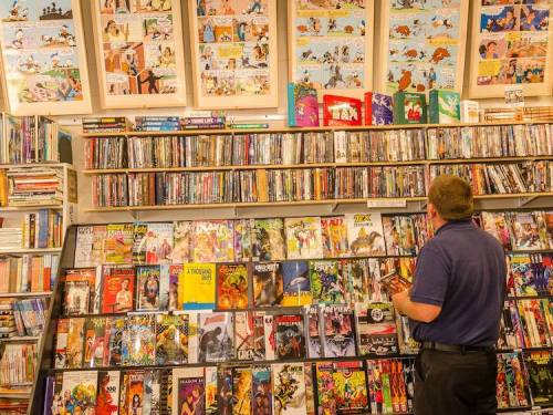 A person browsing a colorful display of comic books and DVDs in a cozy store, with comic art on the walls above.