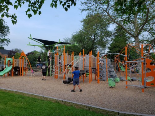 A colorful playground with slides, climbing structures, and children playing on a gravel surface surrounded by trees.