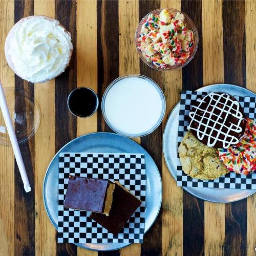 A table with two plates of cookies, a glass of milk, a shot of chocolate syrup, and a dessert cup with sprinkles.