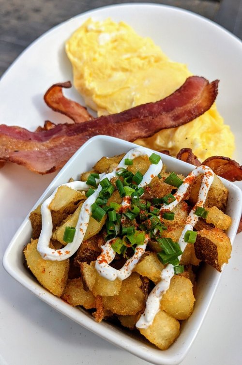 A plate featuring an omelette, crispy bacon, and a bowl of seasoned potato cubes topped with sour cream and chives.