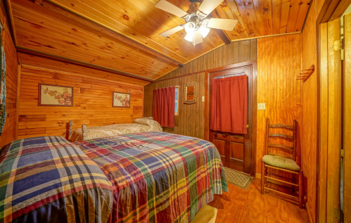 Cozy wooden bedroom with a plaid bedspread, ceiling fan, and red curtains, featuring a door and a chair.