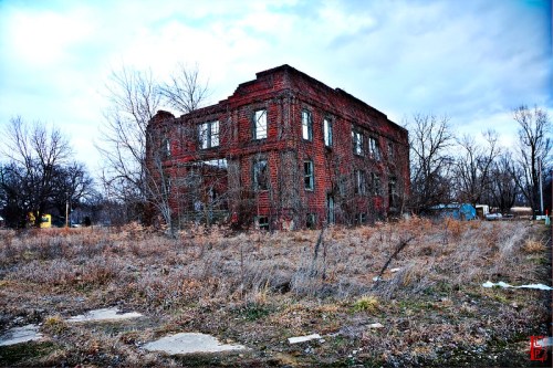 Abandoned brick building surrounded by overgrown grass and trees under a cloudy sky.