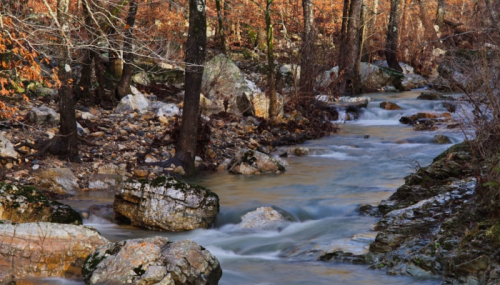 A serene river flows through a rocky landscape, surrounded by trees with autumn leaves.